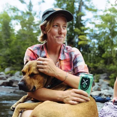 Hannah sits outside with her dog in her lap. She is wearing a baseball cap, has short blonde hair and is looking to the right.