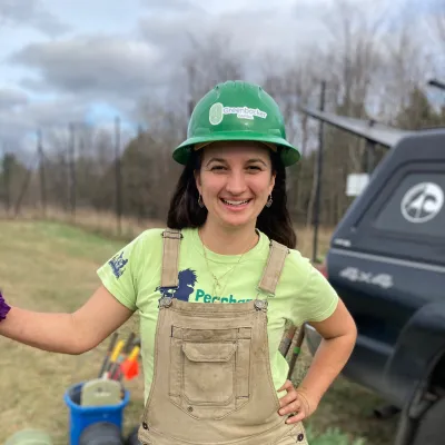 Holly stands outside smiling at the camera. She has a dark green hard hat, light green t-shirt, and brown overalls on. 