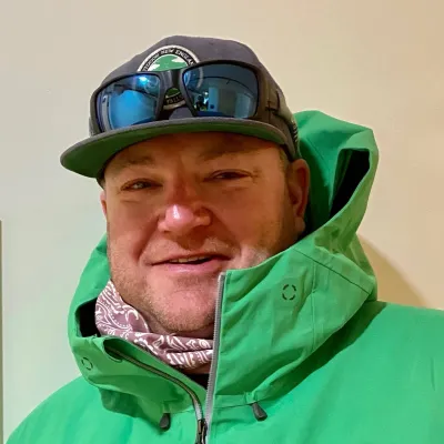 Norm smiles at the camera wearing a green ski jacket and flat brimmed cap with sunglasses perched on top.
