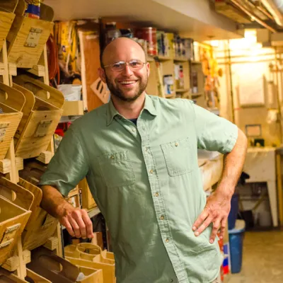 Jacob smiles at the camera in a workshop. He is leaning on a work bench and wearing glasses and a short-sleeved green button-down shirt.