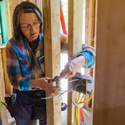 Jan uses pliers to wire a switch on an unfinished house. She has shoulder-length grey hair and is wearing a blue and red flannel shirt.
