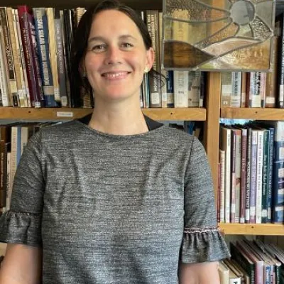 Jen Dirmaier stands in front of bookshelves and smiles at the camera. She has short, dark brown hair and is wearing a grey shirt.