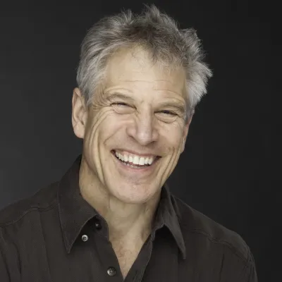John Connell smiles at the camera in front of a black background. He has short, gray hair and is wearing a black collared shirt.