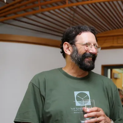 John stands in the main studio, smiling at someone off camera. He is wearing a dark green Yestermorrow shirt, and has dark brown hair and beard