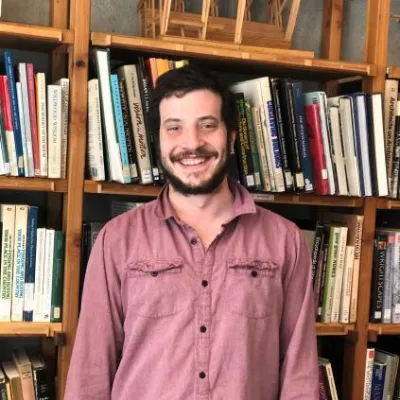 Johno smiles at the camera, standing in front of bookshelves. He is wearing a red button down shirt and has brown hair with a beard.