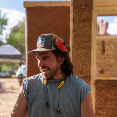 Juan stands in front of an unfinished strawbale cabin. He is wearing a blue hat and t-shirt and orange headphones.