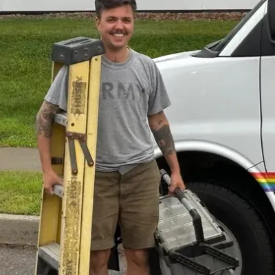 Jules stands outside in front of a white work van with a rainbow stripe. They are carrying a yellow ladder and tool bag.