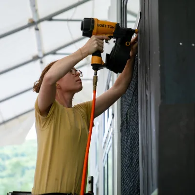 A person with curly red hair and yellow t-shirt uses a nail gun on the black siding of a tiny house.
