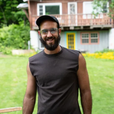 Jussi stands outside in front of a lawn and chalet building. He is wearing a baseball cap and black sleeveless shirt. He has dark brown hair and beard.