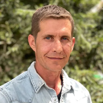 Keith smiles at the camera in a headshot, outside in front of trees. He has short brown hair and is wearing a light blue button-down shirt.