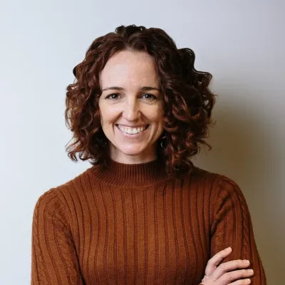 Kelly smiles in a headshot against a white background. She has brown curly hair and is wearing a dark orange sweater with her arms crossed in front.