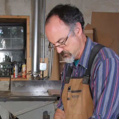Ken pictured in a woodshop at a work table. He has short dark hair and is wearing a blue striped shirt and brown apron.