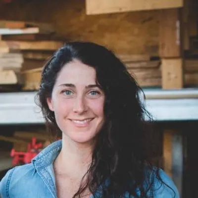 Laura Moss stands in front of a lumber rack, smiling at the camera. She has long, wavy, dark brown hair and is wearing a blue button-down shirt.