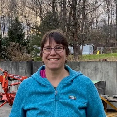 Laura stands outside in front of a jobsite with tools in the back. She has short brown hair and is wearing a bright blue zip up.