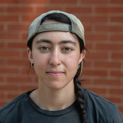 Lily smiles at the camera in front of a brick wall. She has long dark hair in a braid and is wearing a backwards baseball cap and dark blue yestermorrow shirt.