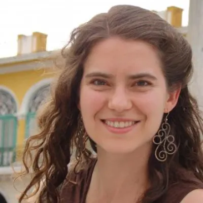 Lina smiles at the camera, outside with a yellow building behind her. She has curly, medium length brown hair and is wearing a brown tank top.