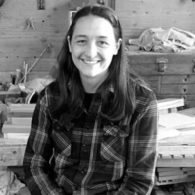 Lindley smiles at the camera in her workshop. She is wearing a plaid shirt and has dark hair. The photo is in black and white.