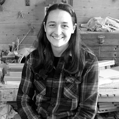 Lindley smiles at the camera in her workshop. She is wearing a plaid shirt and has dark hair. The photo is in black and white.