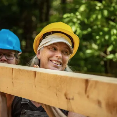 Liz wears a yellow hard hat and is carrying a piece of lumber.