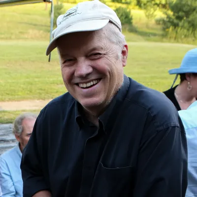 Mac smiles at the camera at an outside event. He is wearing a white baseball cap and black button-down shirt.