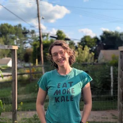 Mackenzie stands outside in front of lawn and sky smiling at the camera. She is wearing jeans and an "eat more kale" shirt, and has curly light brown hair and glasses.