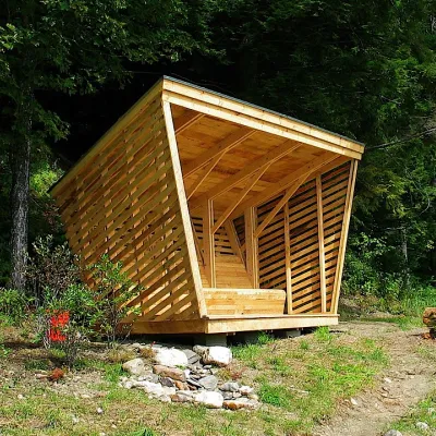 A geometric wooden shelter with slatted walls and a slanted roof stands at the edge of a forest along a dirt trail.