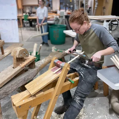 Maret is pictured using a shave horse to make spokes in the Yestermorrow woodshop. They have red hair pulled back and a grey and green long sleeved shirt.