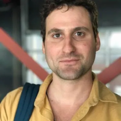 Matt Wolpe smiles at the camera. He has short brown hair, is wearing a gold collared shirt and bag over his shoulder. 