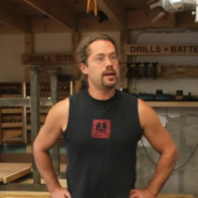 Middle aged man with long hair stands with his hands resting upon his hip.