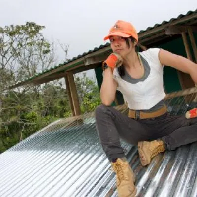 Miriam Gee sits on a metal roofed structure, wearing an orange cap, white shirt, and work pants and boots.