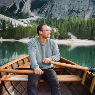 Niel is photographed in a rowboat on a lake with mountains behind him. He has a blue sweater, short brown hair, and is looking off to the right.