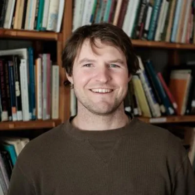 Nick Pattis smiles at the camera standing in front of bookshelves. He has short, light brown hair and is wearing a dark green sweater.