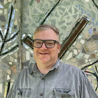 Norm smiles at the camera in front of a tile mosaic wall. He is wearing glasses and a short sleeved blue/gray button down shirt.