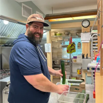 Patrick Dakin smiles at the camera while using a mandolin to cut cucumbers. He has brown hair and a beard and is wearing a brown hat and blue hoodie.