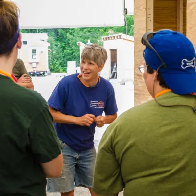 Patti is smiling, talking to a group of students with their backs to the camera. Patti has short gray hair and is wearing a blue t-shirt.