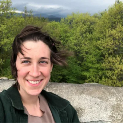 Penny Harte stands on top of a mountain, with trees and sky in the background. She has short, brown hair blowing in the wind and blue eyes and is wearing a green jacket.