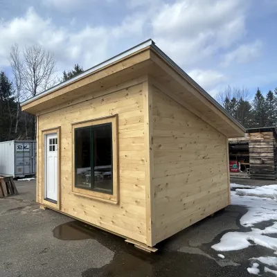 The exterior of a cabin with a large window, white door, pine shiplap siding and a shed roof.