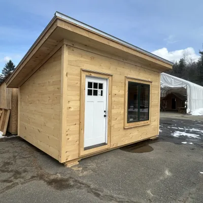 The exterior of a cabin with a large window, white door, pine shiplap siding and a shed roof.