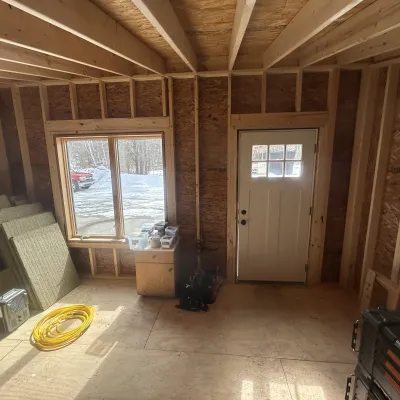 An interior view of a cabin showing exposed wooden wall studs and ceiling joists, with a large window and a white entry door.