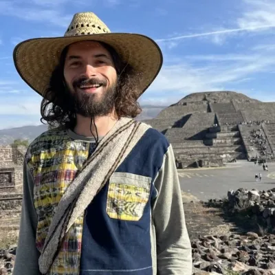 Prem smiles at the camera, photographed outside in a sunny desert scene. He is wearing a wide brimmed hat and a patchwork shirt.