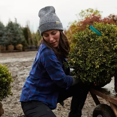 Rachel is outside kneeling next to a wheelbarrow, lifting a plant. She has long brown hair and is wearing a gray beanie and blue flannel shirt.