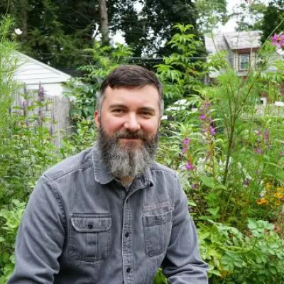 Richard smiles at the camera, outside in a garden. He has short brown hair and long beard and is wearing a gray button-down shirt.