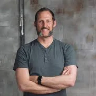 Sam stands in front of a gray wall with his arms crossed. He is wearing a gray t-shirt and has short, light brown hair and beard.