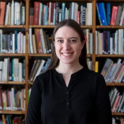 Sarah Johnson, standing in front of bookshelves, smiles at the camera. She has long, brown hair and is wearing a black blouse.