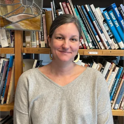 Sarah Wright stands in front of bookshelves and smiles at the camera. She has light brown hair pulled back and is wearing a grey sweater.