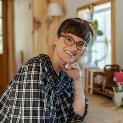 A smiling person with short dark hair and glasses sits at a wooden desk, resting their chin on their hand.
