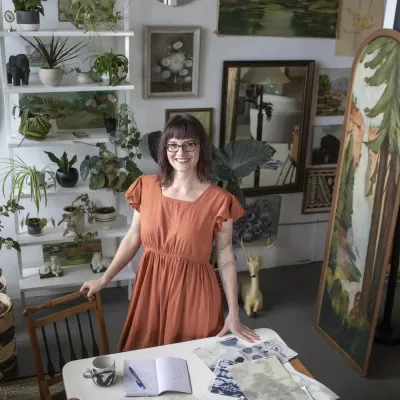 Sarah stands inside in a room filled with plants. She has medium length brown hair and glasses and is wearing a coral dress.
