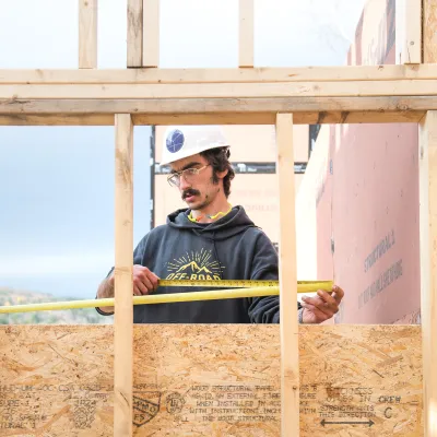 A student uses a tape measure on a partially framed wall. The student is wearing a white hard hat with Yestermorrow logo.