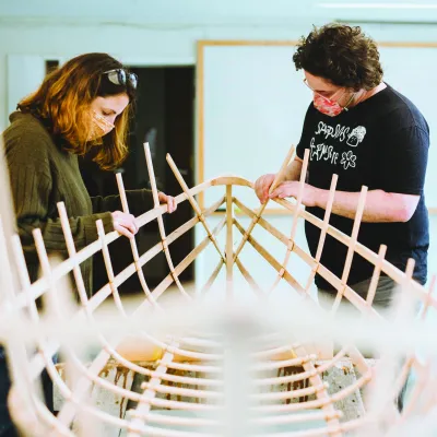 2 people working on the wooden frame of a canoe. 