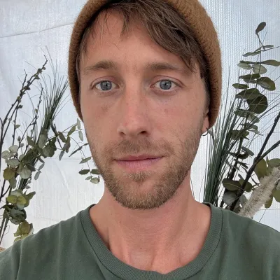 Portrait of a person wearing a brown knit beanie and green shirt, standing in front of a neutral backdrop with natural greenery accents.